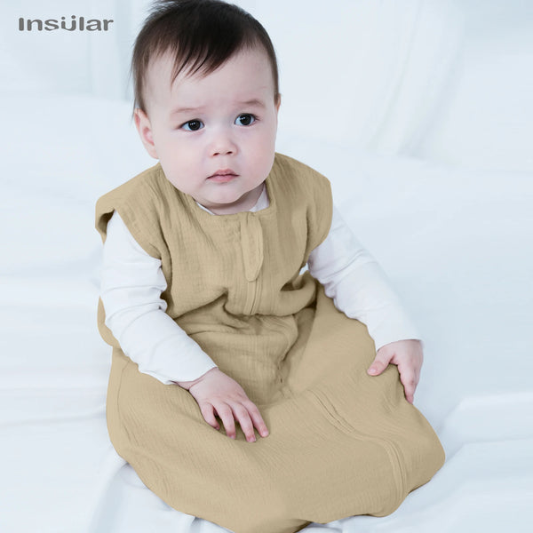 A baby wearing a beige muslin cotton sleeping bag is sitting on a white surface.