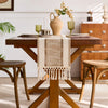 A wooden table set with a beige macrame tassel runner and a vase of dried flowers.