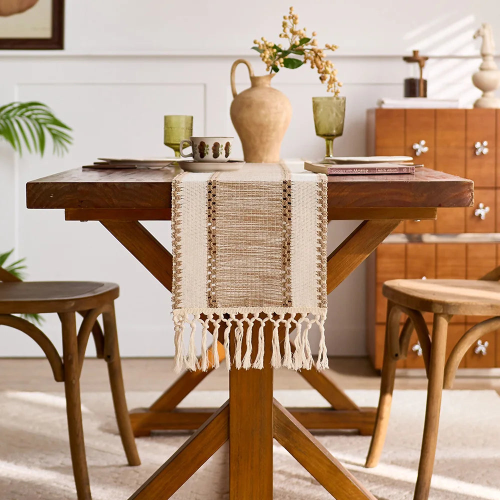 A wooden table set with a beige macrame tassel runner and a vase of dried flowers.