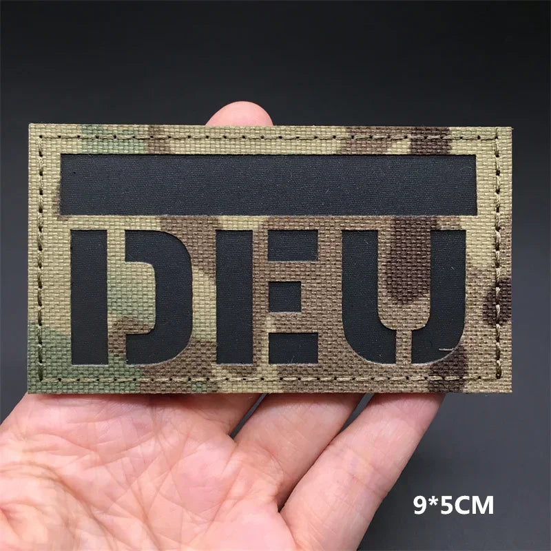 A rectangular tactical flag patch featuring a dark camouflage pattern on a tan fabric, held in a hand.