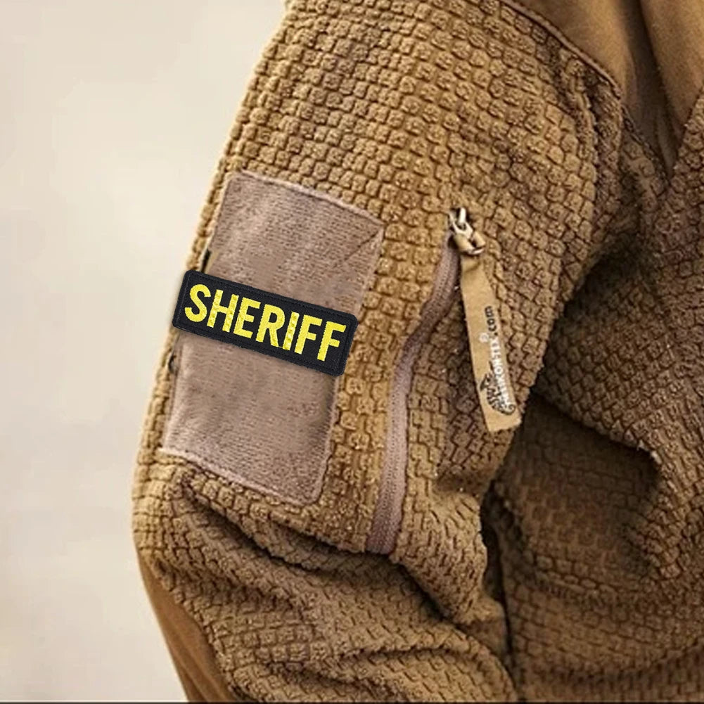 Close-up of a brown tactical jacket sleeve showing a gray patch with the word SHERIFF embroidered on it.