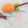An orange crocheted ball is being worked on by a brown crochet hook and various pink and green tools on a gray surface.