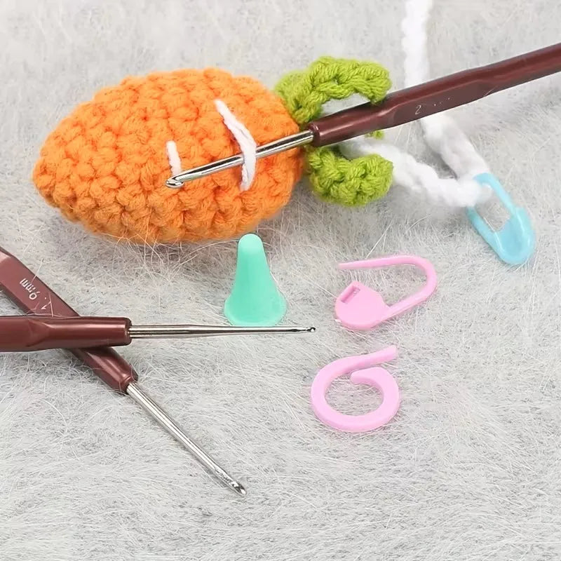 An orange crocheted ball is being worked on by a brown crochet hook and various pink and green tools on a gray surface.