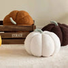 A white fuzzy pumpkin shaped throw pillow is displayed next to two larger, dark orange and brown decorative pumpkins on a woo