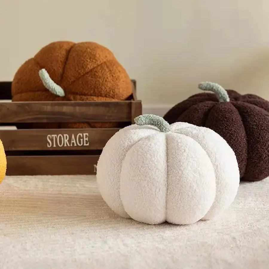 A white fuzzy pumpkin shaped throw pillow is displayed next to two larger, dark orange and brown decorative pumpkins on a woo