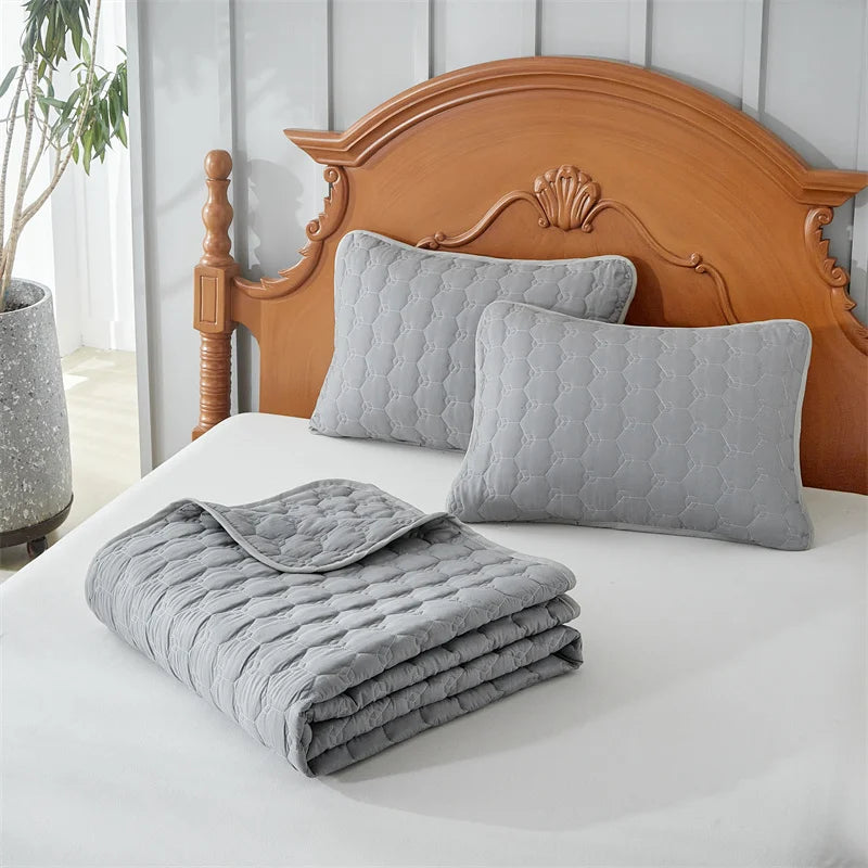 A set of light gray quilted bedspreads and pillows displayed on a white bed with a wooden headboard.