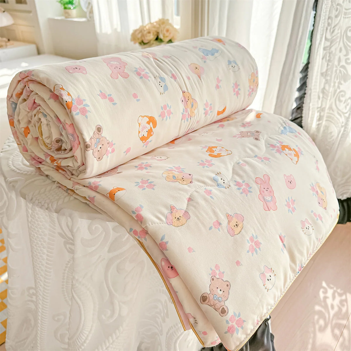 A rolled and unfolded cream-colored quilt blanket with a floral print is displayed on a white bed, next to sheer curtains.