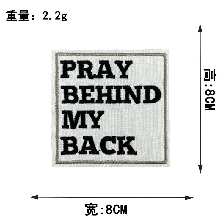A square embroidered patch with black text that reads PRAY BEHIND MY BACK on a light fabric background.
