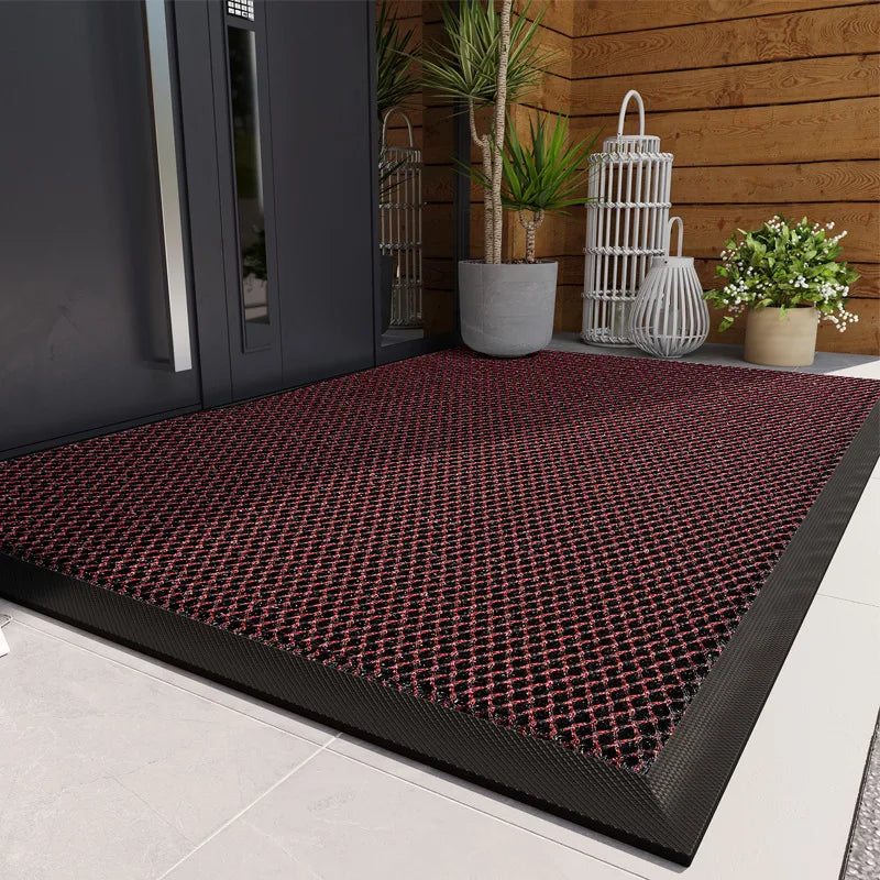 A dark red patterned rubber doormat placed on a light gray floor near a wooden wall and potted plants.