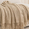 A beige oversized knitted tassel throw blanket draped over a white bed.