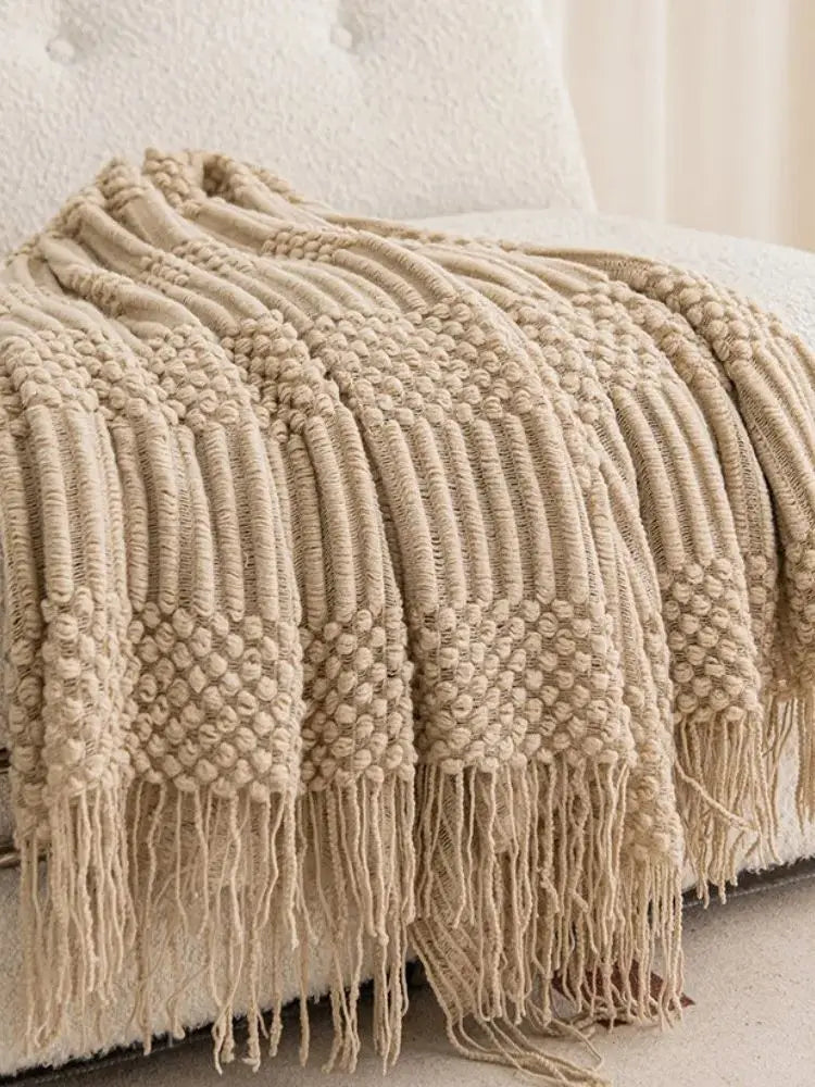 A beige oversized knitted tassel throw blanket draped over a white bed.