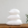 Three white cotton-filled decorative throw pillow inserts stacked on a wooden surface.