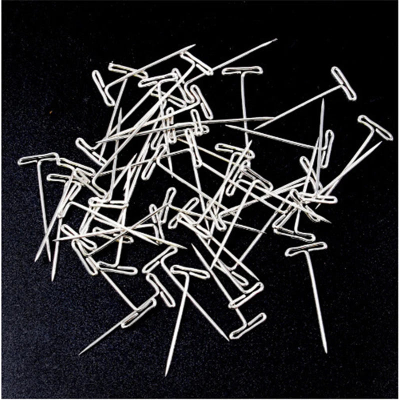 Assorted stainless steel T-pins used for wig making and knitting, scattered on a dark surface.