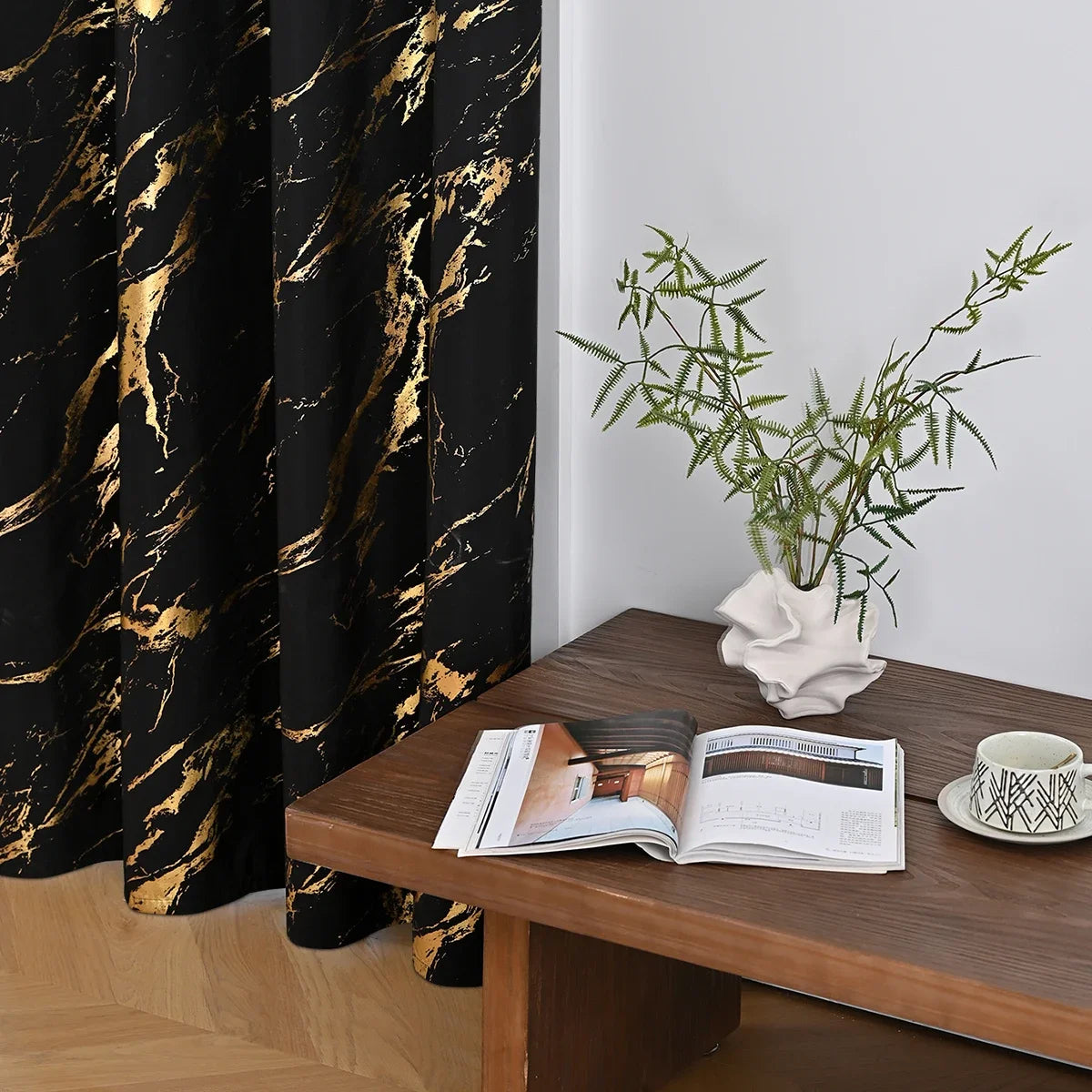Blackout curtains with a gold marble pattern hanging next to an open magazine and a potted green plant on a wooden table.