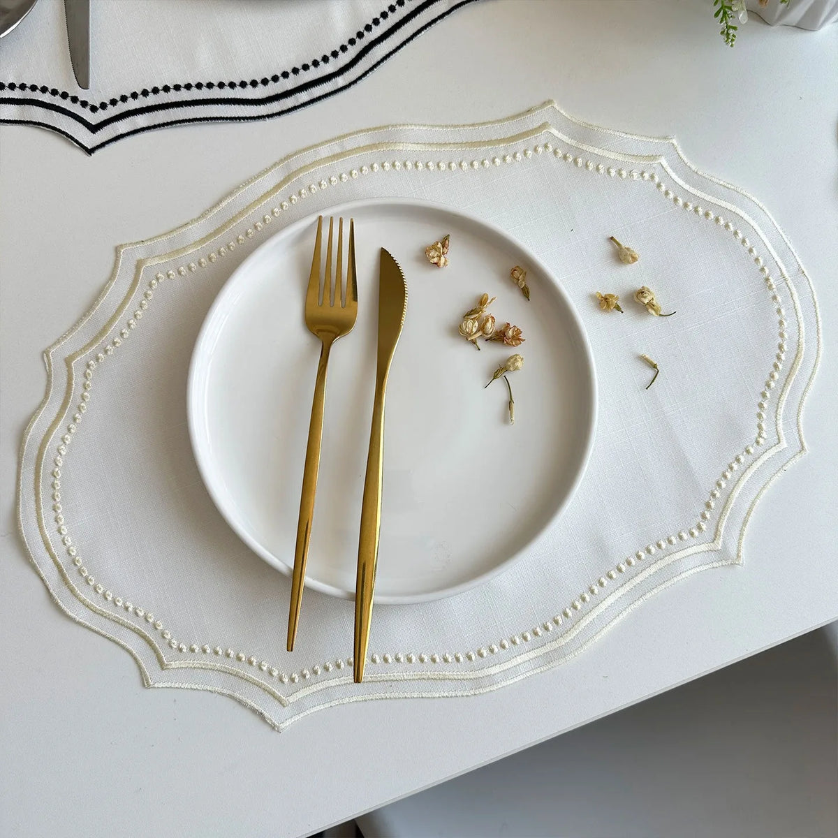A white plate set with gold cutlery is centered on a white linen placemat with delicate embroidery.