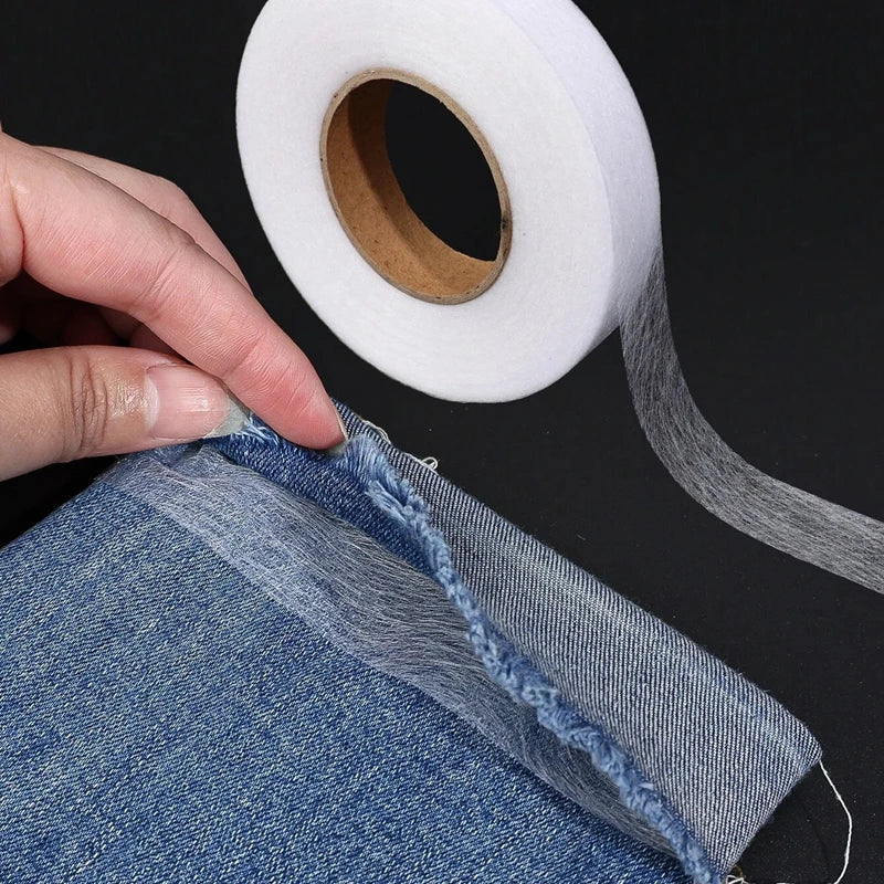 A hand is pulling a piece of denim fabric, showing the edge where heavy duty double-sided iron-on hem tape is applied.