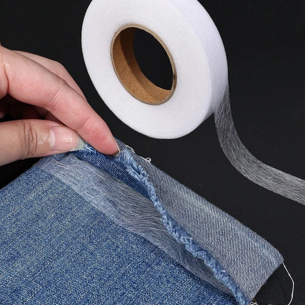 A hand is pulling a piece of denim fabric, showing the edge where heavy duty double-sided iron-on hem tape is applied.