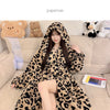 A woman wearing a leopard print flannel kimono robe sitting on a couch surrounded by stuffed animals.
