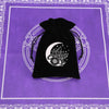 A black velvet pouch with a crescent moon and floral design rests on a purple patterned surface.