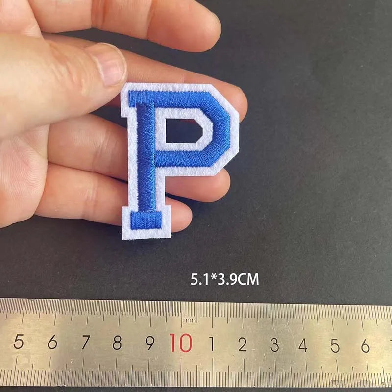 A hand holds a blue alphabet embroidery patch shaped like the letter P, measured against a ruler.