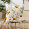 A cream colored throw pillow cover embroidered with white and yellow daisies on a neutral beige knitted blanket.
