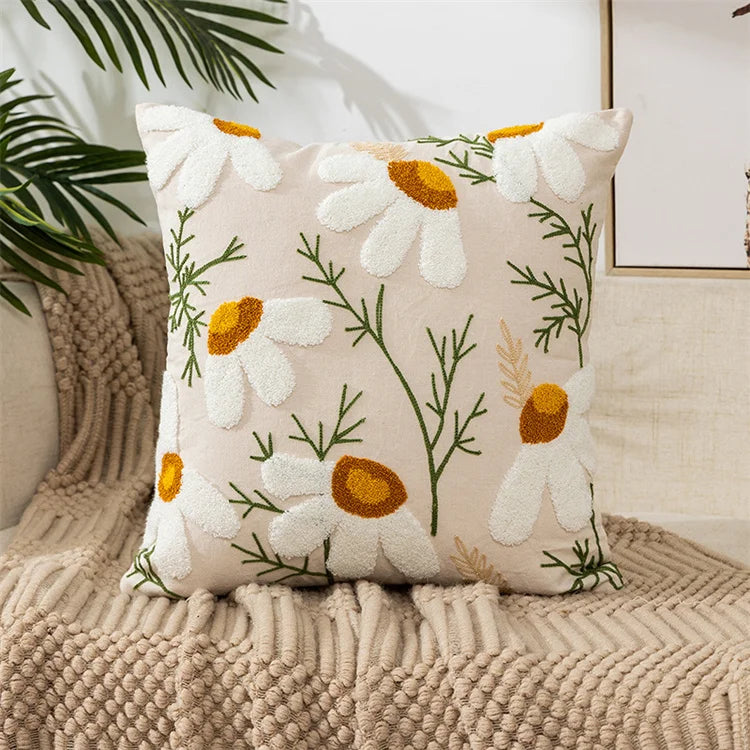 A cream colored throw pillow cover embroidered with white and yellow daisies on a neutral beige knitted blanket.