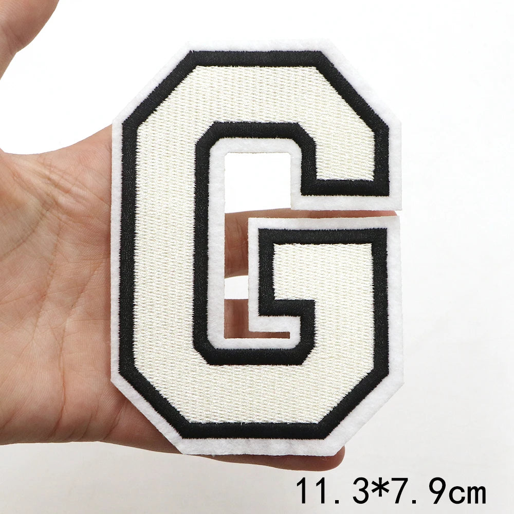 A white letter G patch with a black outline is held in a hand.