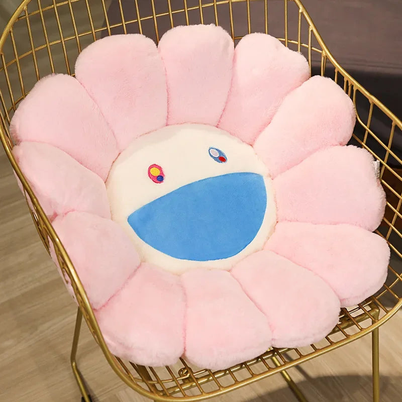 A pink sunflower plush cushion with a white face and blue mouth is displayed in a gold wire basket.