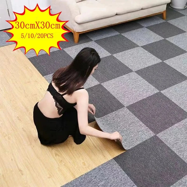 A woman is laying down a square carpet tile on a light wood floor next to a gray and white checkered rug.