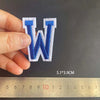 A hand holds a blue alphabet embroidery patch shaped like the letter W against a ruler.
