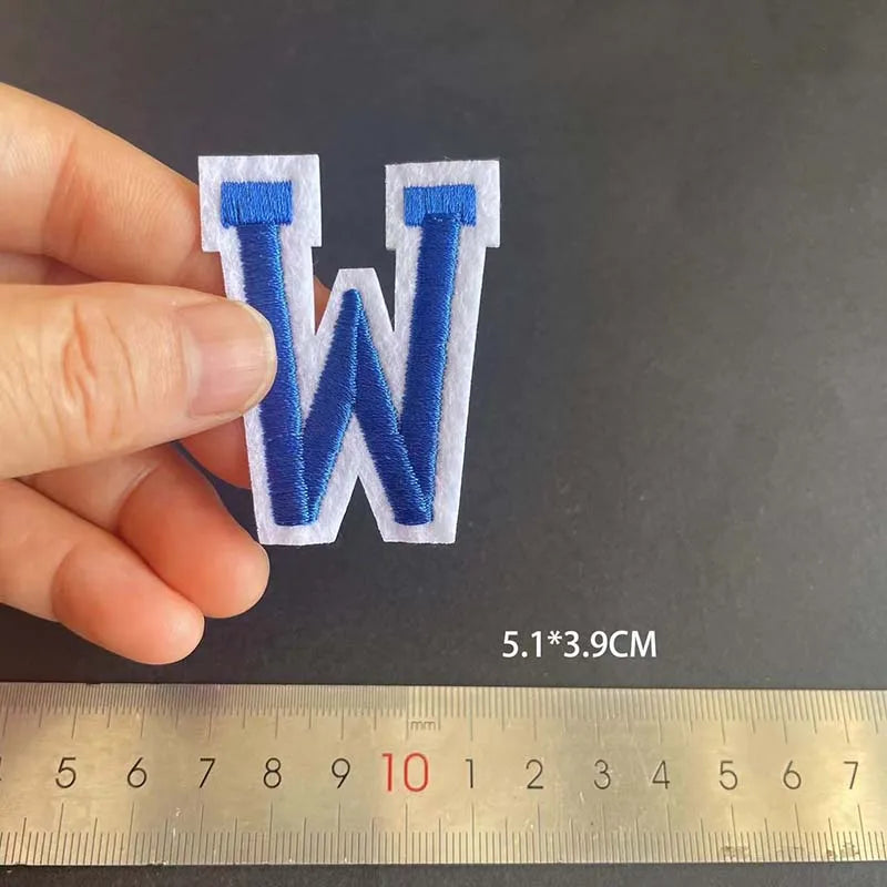 A hand holds a blue alphabet embroidery patch shaped like the letter W against a ruler.
