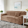 A tan stretch velvet sofa slipcover with a throw pillow on a light gray fluffy rug in a bright room.