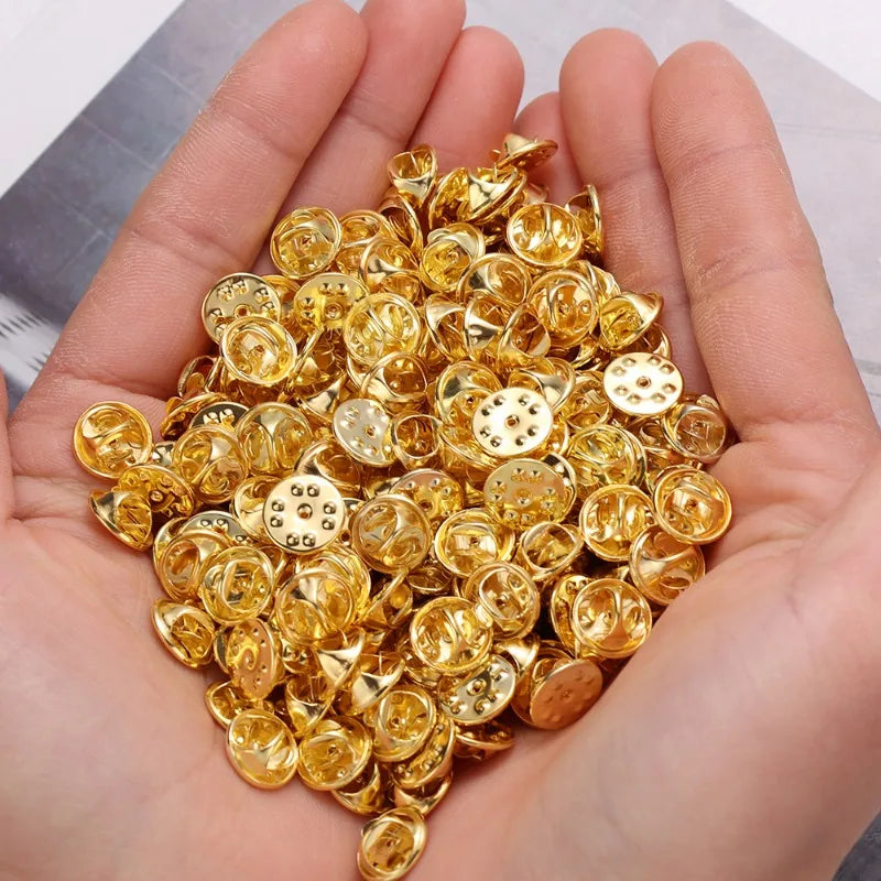 A hand holds a pile of gold-colored metal butterfly brooch clasps and tie tacks for jewelry making.