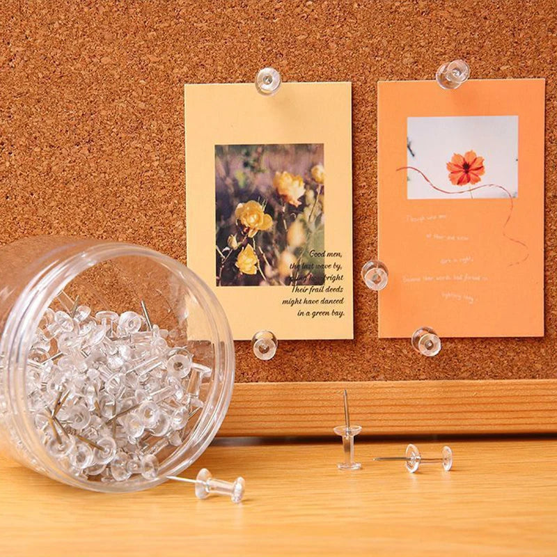 A clear plastic container filled with silver push pins and steel tips resting on a wooden surface.