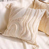 A beige plush pillow cover with wavy geometric lines and tassel details resting on a light fabric.
