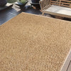 A close-up view of a natural sisal jute rug placed on a dark wooden deck with outdoor furniture in the background.