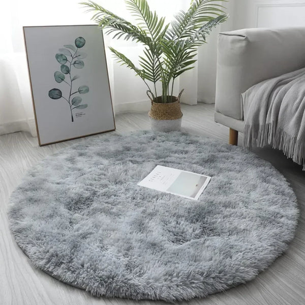 A fluffy gray shaggy circle area rug placed on a light wooden floor with a potted plant and framed artwork in the background.