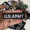 A hand holds several black iron-on slogan patches featuring various military and patriotic themes.