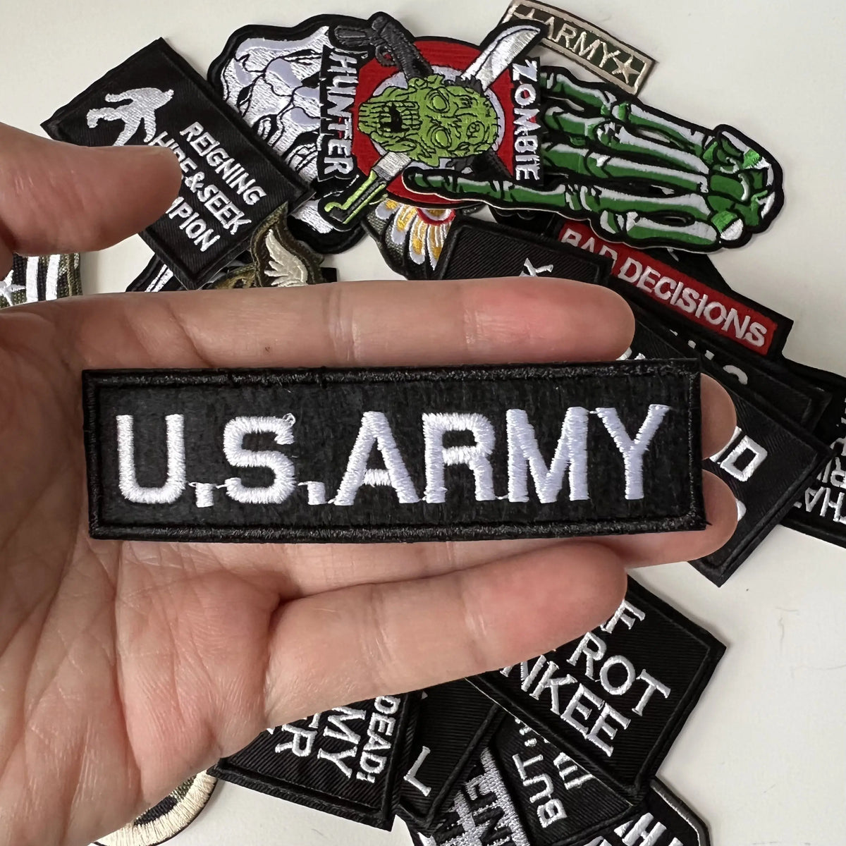 A hand holds several black iron-on slogan patches featuring various military and patriotic themes.
