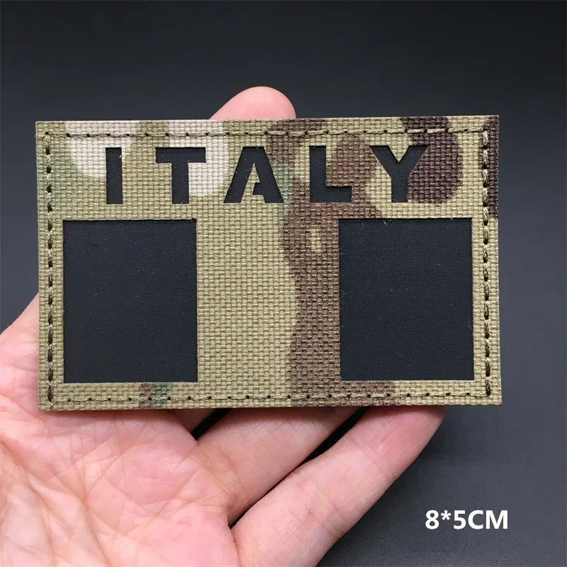 A hand holds a rectangular tactical patch featuring the word ITALY in camouflage pattern.