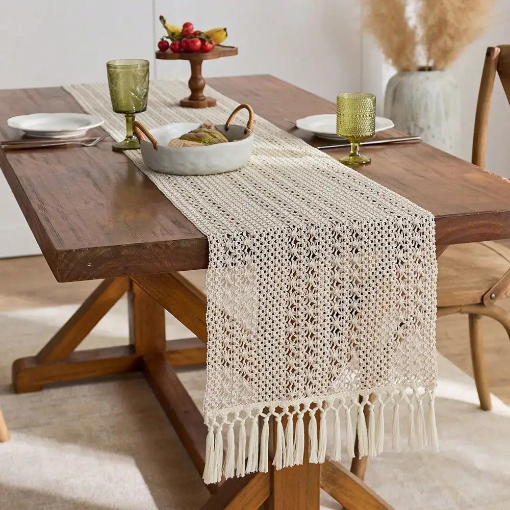 A rustic wooden table is set with a beige macrame tasseled table runner, small bowls of fruit, and gold goblets.