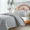 A light gray quilted bedspread set is draped over a white bed in a bedroom setting.