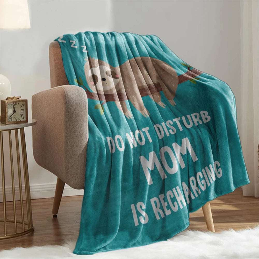 A teal knit throw blanket featuring a sloth design with the text DO NOT DISTURB MOM IS RECHARGING