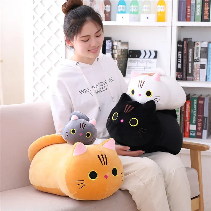A woman is sitting on a couch with several soft cat plush toys, including a large yellow and black cat, a white cat, and a bl
