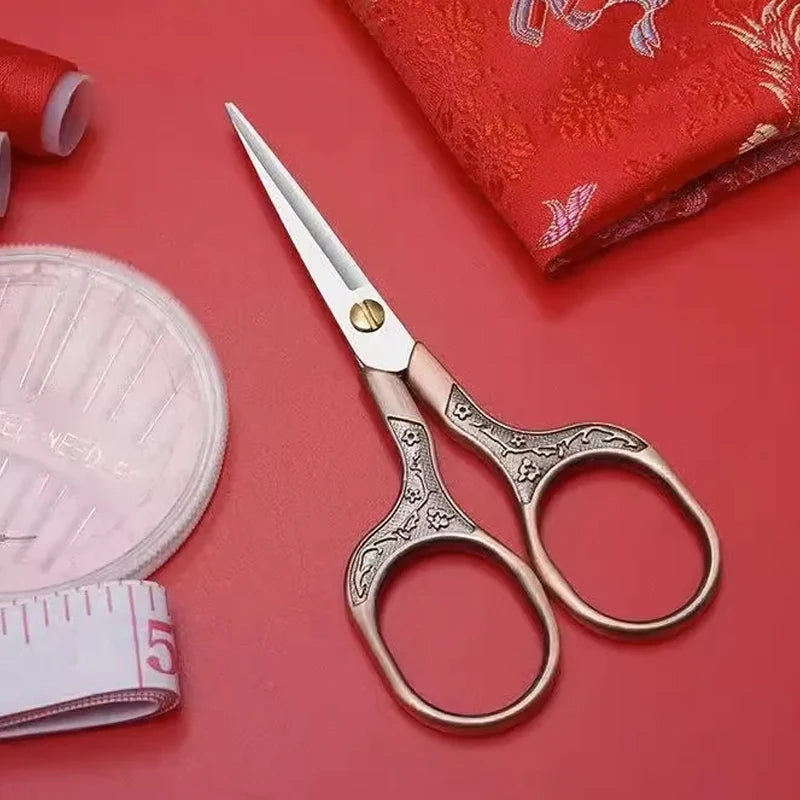 A pair of vintage stainless steel sewing scissors rests on a red fabric background next to a pink measuring tape and a rolled