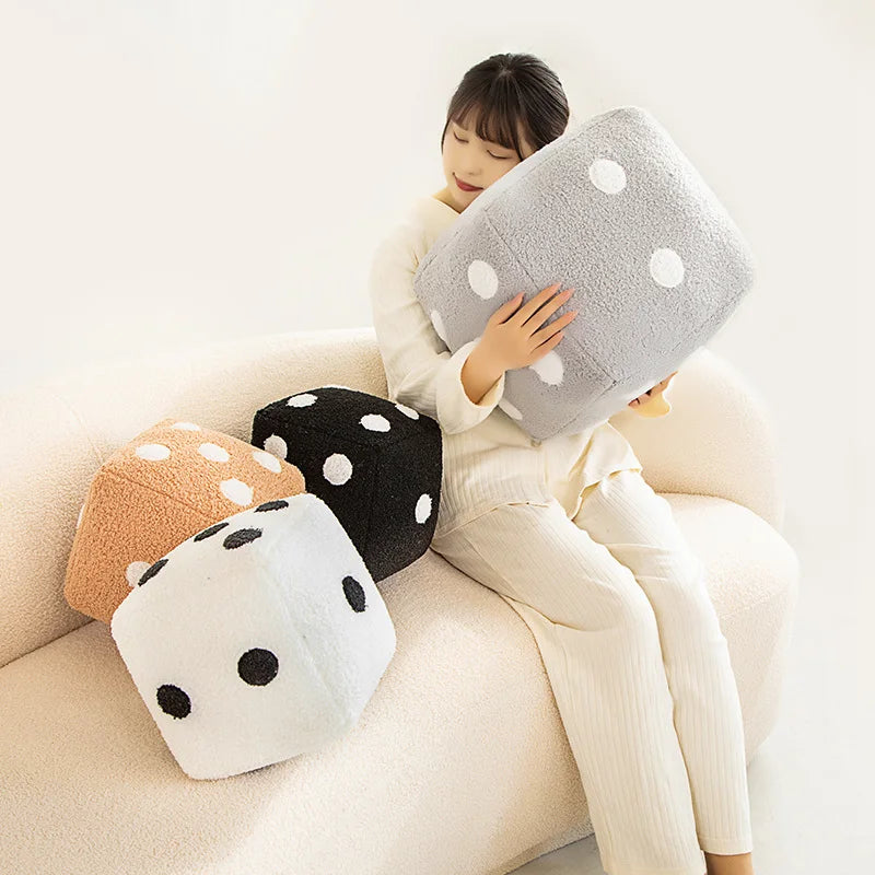 A woman is holding a gray plush dice cube cushion and several black and white polka dot cushions on a light-colored sofa.