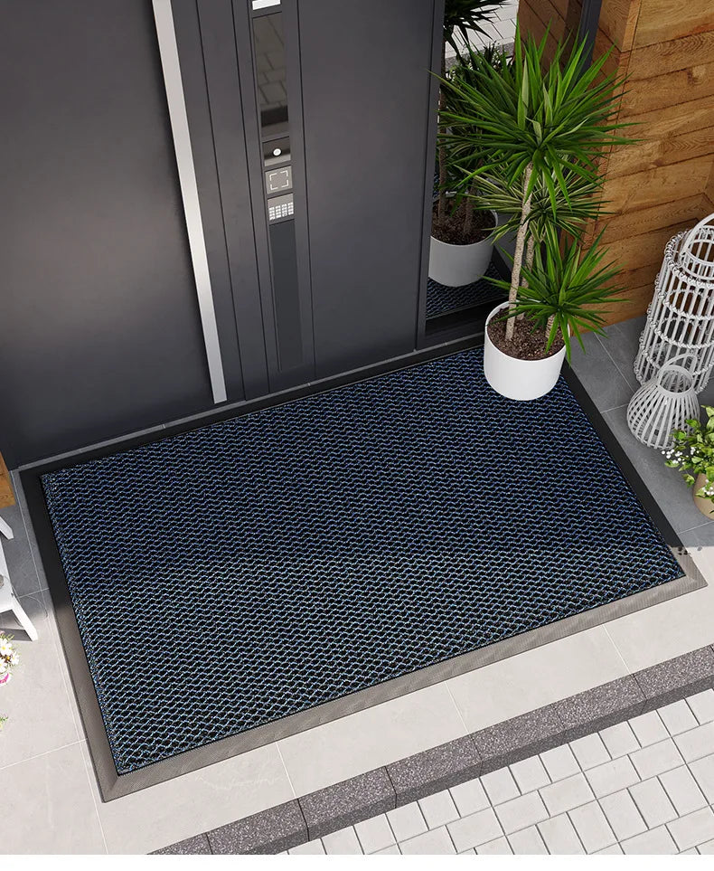 A dark blue patterned non-slip rubber doormat placed on a gray tiled floor near a modern gray door and potted plant.