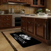 A black non-slip flannel area rug is placed on a light wood floor in a kitchen setting.