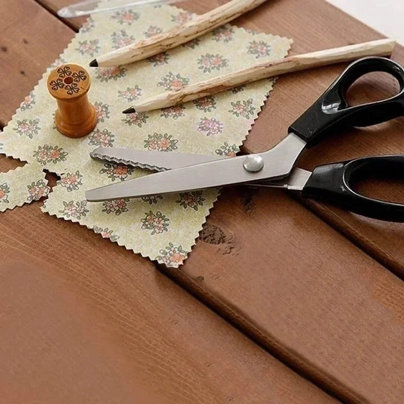 A pair of professional dressmaking pinking shears rests on a wooden surface with pencils and patterned paper.