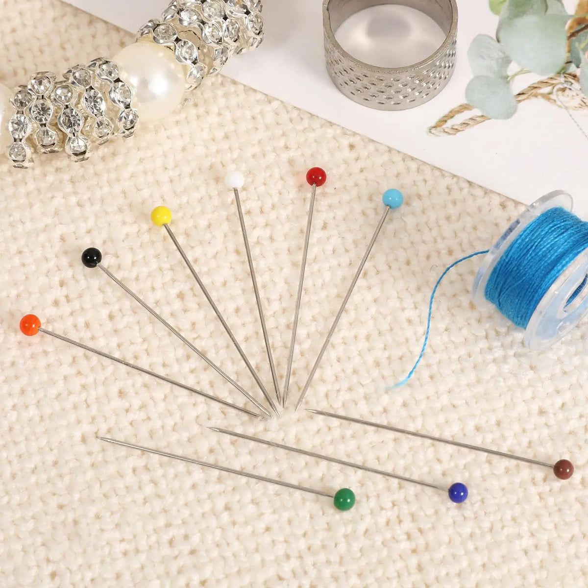 A collection of glass head straight pins in various colors and sizes laid out on a beige fabric surface.
